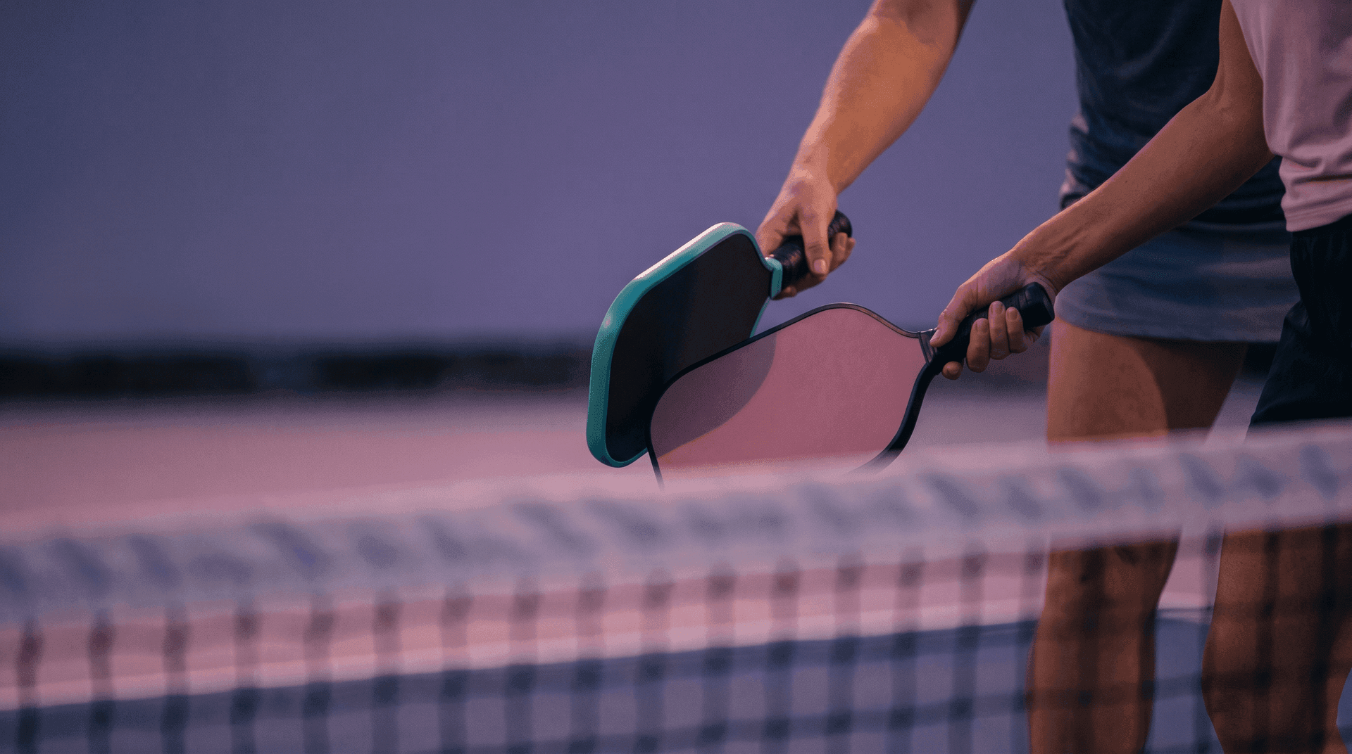 Close-up of two pickleball paddles tapped together at the net in the "good game" gesture, dusk light, no faces visible.